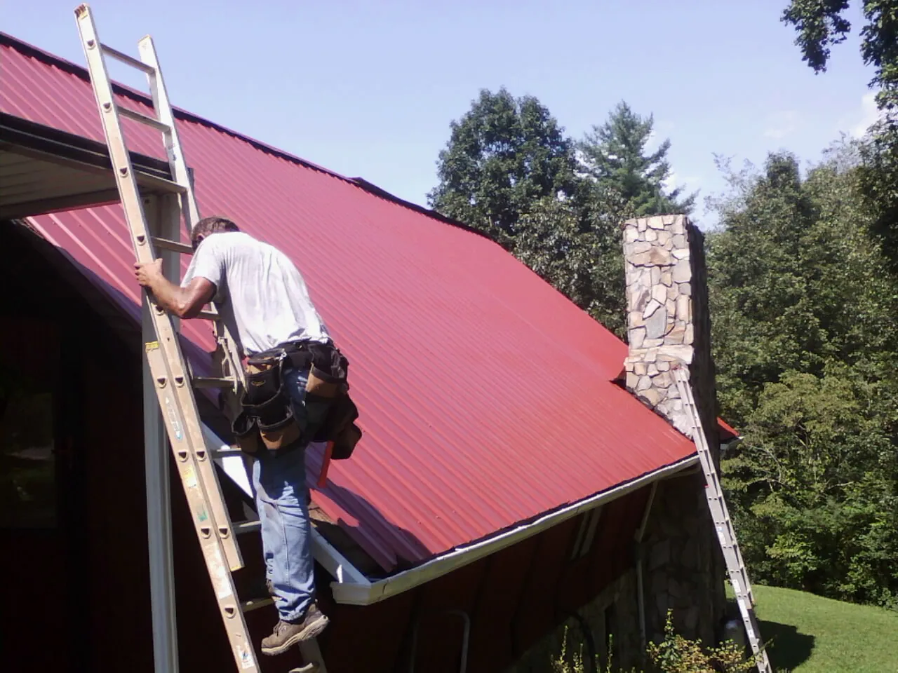 Roofer on a ladder beside a red metal roof and stone chimney — Soffit & Fascia Repair by Roofing Brothers in Norway